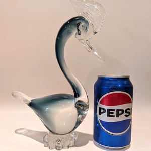May include: A clear glass swan sculpture with a blue-tinted body and a clear, textured crest. The swan sits on a clear, textured base. A blue Pepsi can is next to the sculpture.