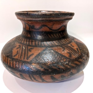 May include: A small, round, terracotta-colored earthenware pot with a wide, flared rim. The exterior surface is decorated with hand-painted black geometric patterns, including triangles, cross-hatching, and wavy lines encircling the neck.