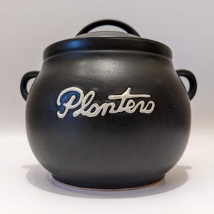 Op de afbeelding: Een zwarte keramische pot met een deksel en twee handvatten aan de zijkant. Het woord "Planters" staat in witte sierletters op de voorkant van de pot. De pot heeft een ronde vorm en staat op een wit oppervlak.