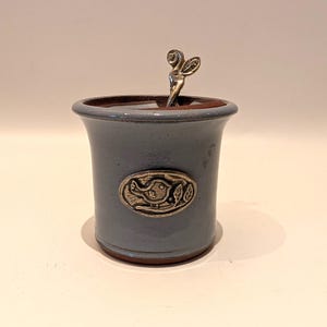 May include: A blue ceramic jar with a silver spoon. The jar has a decorative metal emblem with a bird and leaf design. The rim is brown. The spoon has a decorative handle.