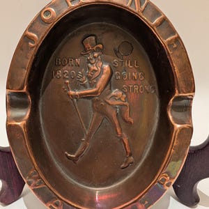 Op de afbeelding: Ovale koperen asbak met de woorden "JOHNNIE WALKER" rond de rand. Het midden toont een verhoogd reli&euml;f van een man met hoge hoed en wandelstok. Tekst luidt "BORN 1820 STILL GOING STRONG."