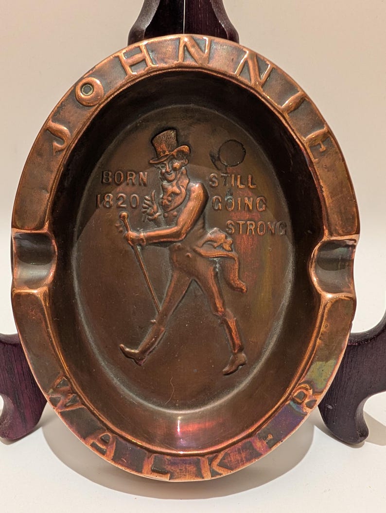 Op de afbeelding: Ovale koperen asbak met de woorden "JOHNNIE WALKER" rond de rand. Het midden toont een verhoogd reli&euml;f van een man met hoge hoed en wandelstok. Tekst luidt "BORN 1820 STILL GOING STRONG."
