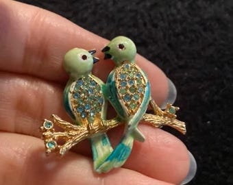 May include: A decorative brooch featuring two birds perched on a golden branch. The birds have light green heads, teal and blue bodies, and are adorned with blue and green gemstones. The branch has small blue gemstones.