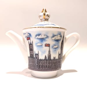 Może przedstawiać: Biały porcelanowy imbryk ze szczegółową czarno-białą ilustracją Pałacu Westminsterskiego i Big Bena, ozdobiony niebieskimi chmurami i małymi flagami Union Jack. Pokrywka zwieńczona jest złotym uchwytem w kształcie korony.