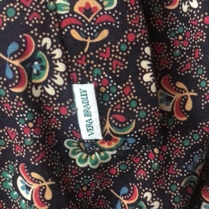 K&ouml;nnte beinhalten: Nahaufnahme eines dunkelblauen Stoffes mit einem farbenfrohen Blumenmuster in Rot, Gelb, Gr&uuml;n und Blau. Der Stoff hat ein kleines wei&szlig;es Etikett mit der Aufschrift "VERA BRADLEY". Das Muster umfasst Paisley- und Blumendesigns.