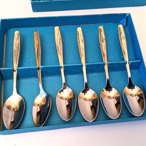 May include: A set of six gold-coloured spoons with a decorative design on the handles, presented in a blue box. The spoons have a polished, reflective surface. The spoons are arranged neatly in individual slots within the box.