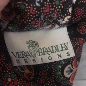 K&ouml;nnte beinhalten: Nahaufnahme eines Stoffes mit einem Muster aus roten, gr&uuml;nen und gelben Punkten auf dunklem Hintergrund. Ein wei&szlig;es Etikett mit dem Text "VERA BRADLEY DESIGNS" ist sichtbar.