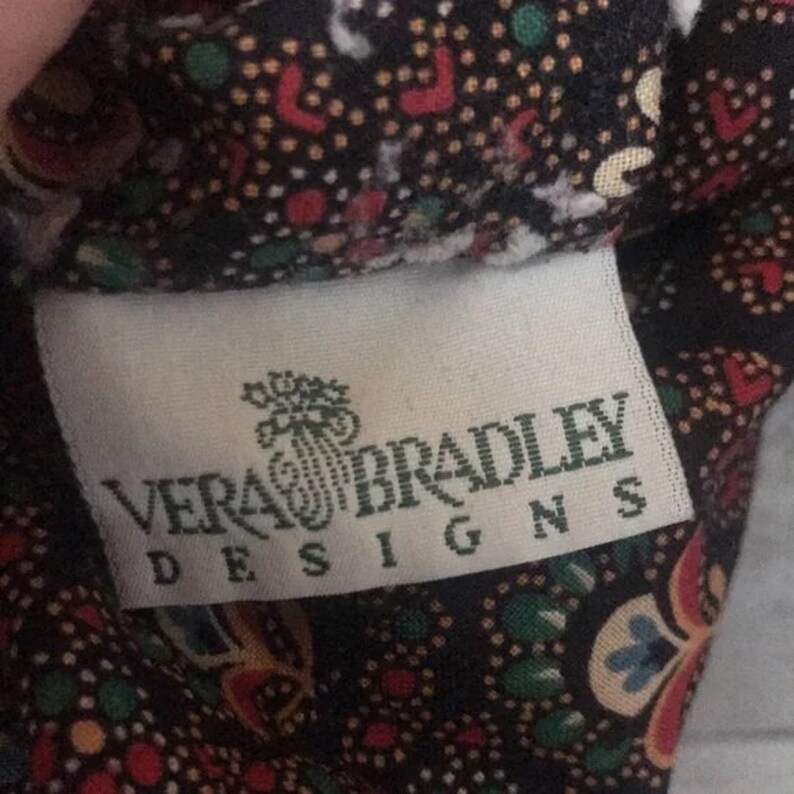 K&ouml;nnte beinhalten: Nahaufnahme eines Stoffes mit einem Muster aus roten, gr&uuml;nen und gelben Punkten auf dunklem Hintergrund. Ein wei&szlig;es Etikett mit dem Text "VERA BRADLEY DESIGNS" ist sichtbar.