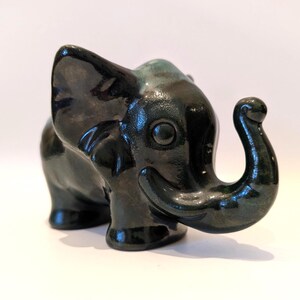 May include: A ceramic elephant figurine in a glossy, dark green glaze. The elephant has a smiling expression, large ears, and a curled trunk. The figurine is standing on a white surface.