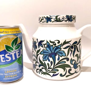 Könnte beinhalten: Weiße Keramik-Teekanne mit Deckel, verziert mit blauen und grünen Blumenmustern. Die Teekanne hat einen gebogenen Griff und Ausguss. Eine Dose Nestea Lemon Iced Tea steht neben der Teekanne.