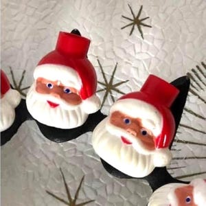 Könnte beinhalten: Ein kreisförmiger Kerzenhalterrahmen aus schwarzem Metall mit mehreren geformten Kunststoff-Weihnachtsmannköpfen. Jede Figur hat eine leuchtend rote Mütze, einen weißen Bart und bemalte Gesichtszüge, einschließlich blauer Augen.