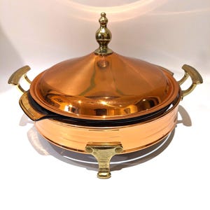 Puede incluir: Una fuente de servir de color cobre con tapa y asas y patas de latón. La tapa tiene un adorno decorativo de latón. La fuente redonda tiene un interior negro y está diseñada para servir alimentos.