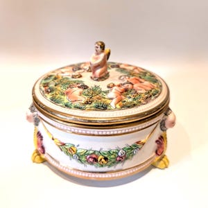 May include: A round, white ceramic trinket box with a lid depicting cherubs and foliage. The box has gold trim, floral garland accents, and small feet. A small cherub figure sits on the lid.