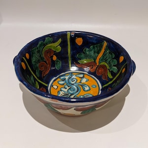 May include: A blue ceramic bowl with a colourful floral design. The bowl has two small handles on the sides and is decorated with yellow, orange, green, and brown accents.