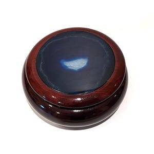 May include: A circular wooden box with a polished, dark reddish-brown exterior. The lid features a dark blue agate stone centre with a lighter blue and white pattern. The box has a glossy finish.