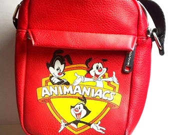 May include: A bright red crossbody bag with a textured finish featuring a graphic print of the Animaniacs characters Yakko, Wakko, and Dot inside a yellow shield emblem. The bag includes a front zippered pocket and a black adjustable shoulder strap.