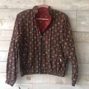 K&ouml;nnte beinhalten: Wende-Bomberjacke mit braunem Blumenmuster auf der einen Seite und rotem Blumenmuster auf der anderen Seite. Die Jacke hat einen schwarzen Rei&szlig;verschluss, einen Stehkragen und elastische B&uuml;ndchen und Saum. Die Jacke h&auml;ngt an einem rosa Kleiderb&uuml;gel.