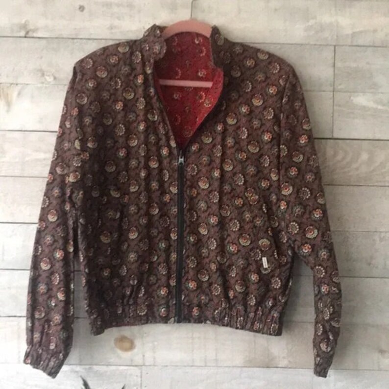 K&ouml;nnte beinhalten: Wende-Bomberjacke mit braunem Blumenmuster auf der einen Seite und rotem Blumenmuster auf der anderen Seite. Die Jacke hat einen schwarzen Rei&szlig;verschluss, einen Stehkragen und elastische B&uuml;ndchen und Saum. Die Jacke h&auml;ngt an einem rosa Kleiderb&uuml;gel.