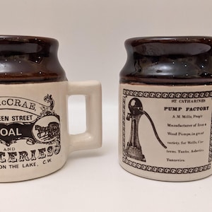 May include: Two brown and white ceramic mugs with handles. The mug on the left has the text "D. McCRAE, 104 QUEEN STREET, COAL AND GROCERIES, NIAGARA ON THE LAKE, C.W." The mug on the right has the text "ST. CATHARINES PUMP FACTORY, A.M. Mills, Prop'r, Manufacturer of Iron & Wood Pumps, in great variety, for Wells, Cisterns, Tanks, Advertisers, Tanneries."