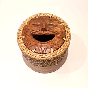 Könnte beinhalten: Ein runder Holzbehälter mit einem geschnitzten Deckel, der ein lächelndes Sonnengesicht zeigt. Der Deckel ist von einem geflochtenen Rand aus hellem Schilf oder Weide umgeben, der mit dem glatten, zylindrischen Holzsockel des kleinen Gefäßes verbunden ist.