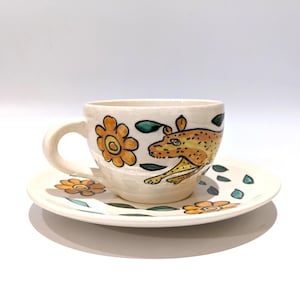 Puede incluir: Un juego de taza y plato de cerámica con un diseño pintado a mano de un leopardo amarillo con manchas y flores naranjas con hojas verdes. La taza tiene un asa curva y el plato combina con el motivo floral y animal en un acabado esmaltado crema.
