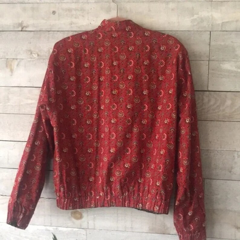K&ouml;nnte beinhalten: Rote Jacke mit floralem Muster. Die Jacke hat lange &Auml;rmel, einen Stehkragen und einen gerippten Saum. Das florale Muster enth&auml;lt wei&szlig;e, beige und schwarze Akzente. Die Jacke h&auml;ngt an einem Metallb&uuml;gel.