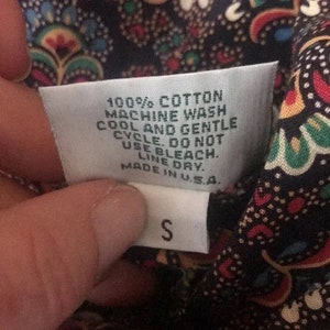 K&ouml;nnte beinhalten: Nahaufnahme eines wei&szlig;en Kleidungsetiketts mit dem Text "100% COTTON MACHINE WASH COOL AND GENTLE CYCLE. DO NOT USE BLEACH. LINE DRY. MADE IN U.S.A." und dem Buchstaben "S". Das Etikett ist an einem dunklen Stoff mit Paisley-Muster befestigt.