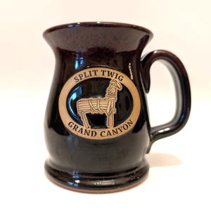 Peut inclure: Une tasse en céramique marron foncé avec une anse. La tasse présente un emblème ovale avec les mots "Split Twig Grand Canyon" et une image stylisée d'un mulet. La tasse a une finition brillante.