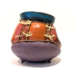 Peut inclure: Un petit récipient en céramique arrondi avec un bord bleu sarcelle foncé, une section médiane lisse de couleur terre cuite et une base violette texturée. Le milieu est entouré d'une grille de brindilles brunes fixées par des liens en raphia.