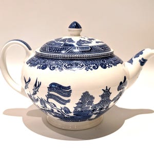 Può includere: Una teiera in ceramica bianca con un motivo blu in stile willow. Il design presenta illustrazioni dettagliate di pagode, salici e uccelli. È dotata di un manico curvo, un beccuccio e un coperchio abbinato con un piccolo pomello sulla parte superiore.
