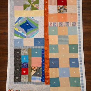 May include: A rectangular patchwork quilt featuring a variety of fabric squares in shades of blue, green, orange, red, and floral patterns. The quilt is divided by a vertical orange strip and includes white hand-stitched embroidery details throughout.