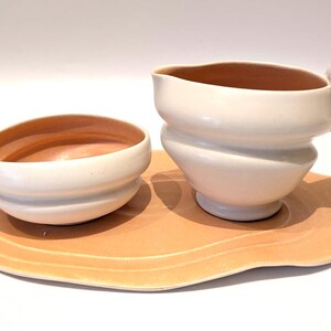May include: A cream-colored ceramic creamer and bowl set with a peach-colored interior, resting on a matching peach-colored, organically shaped tray. The creamer has a handle and a spout.