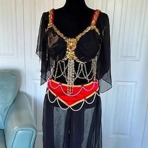 Könnte beinhalten: Schwarzes Bauchtanzkostüm mit goldenen und roten Akzenten. Das Oberteil hat goldene Verzierungen und einen roten Gürtel. Das Kostüm beinhaltet einen schwarzen Rock und transparente Ärmel. Das Outfit wird auf einer schwarzen Schaufensterpuppe präsentiert.