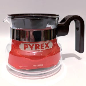 May include: A clear Pyrex glass coffee pot with a dark brown handle and lid. The pot has a red label with the Pyrex logo and the text "0.5 litre". The pot is made in France.