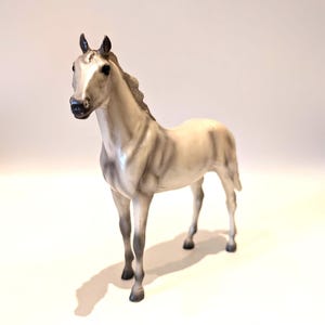 Könnte beinhalten: Detaillierte Figur eines Pferdes in stehender Pose. Das Pferd ist überwiegend weiß mit grauen Schattierungen und schwarzen Hufen, Mähne und Ohren. Die Figur ist eine realistische Darstellung eines jungen Pferdes.