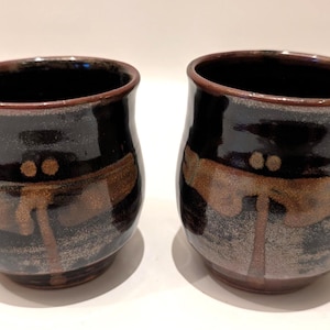 Op de afbeelding: Twee keramische bekers met een donkerbruin glazuur en een libellenontwerp in bruin- en beigetinten. De bekers hebben een afgeronde vorm met een licht uitlopende rand. De binnenkant van de bekers is zwart.