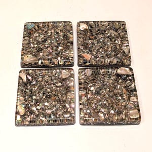 May include: A set of four square coasters featuring a mosaic of crushed iridescent abalone shells embedded in a clear resin base. Each coaster has a circular recessed center and a smooth, glossy finish with varied gray, brown, and pearlescent tones.