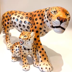 Könnte beinhalten: Eine Keramikfigur eines großen erwachsenen Leoparden, der über einem kleineren Leopardenjungen steht. Beide Tiere haben ein glänzendes orange-braunes Fell mit markanten schwarzen Flecken. Der erwachsene Leopard schaut nach vorne, das Junge hat das Maul offen.