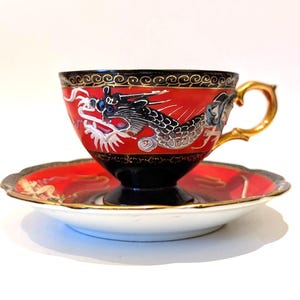May include: A red and black teacup and saucer set with a gold handle and rim. The teacup features a detailed dragon design in black, white, and silver. The saucer has a matching red and gold design. A decorative tea set.
