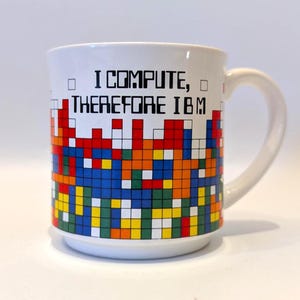 May include: White ceramic mug with a pixelated design in red, blue, yellow, green, and orange. The mug features the text "I COMPUTE, THEREFORE IBM" in black, with a white handle.