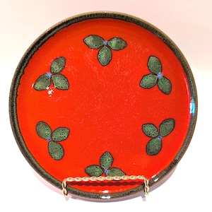 May include: A round ceramic plate with a vibrant orange glazed surface, featuring five green leaf-shaped motifs arranged in a circular pattern. The plate has a dark, textured rim and is displayed on a gold-toned wire stand.