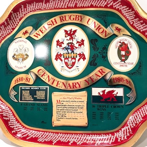 Puede incluir: Bandeja decorativa con el escudo de la Welsh Rugby Union y texto. La bandeja conmemora el Centenario 1880-81 e incluye listas de clubes y logros. Borde rojo con texto blanco y fondo verde.