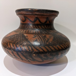 May include: A brown ceramic vase with a rounded body and a narrow neck. The vase is decorated with black geometric patterns, including zigzags and lines. The vase has a diameter of approximately 7 inches.