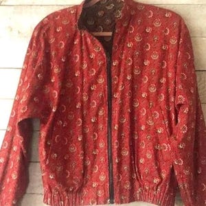 K&ouml;nnte beinhalten: Eine rote Bomberjacke mit schwarzem Rei&szlig;verschluss und Paisley-Muster. Die Jacke ist wendbar, mit einem braunen Paisley-Muster auf der Innenseite. Die Jacke h&auml;ngt an einem Holzkleiderb&uuml;gel.