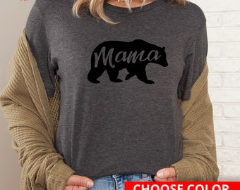 May include: A dark grey t-shirt with a black bear silhouette and the word "Mama" printed on it. The shirt is being worn by a person with long blonde hair. The text "Choose Color & Size (returns accepted)" is printed on a red rectangle at the bottom of the image.