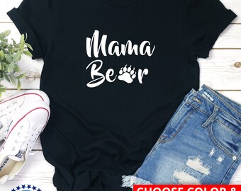 May include: Black t-shirt with white text that says "Mama Bear" with a white paw print. The text is in a cursive font. The t-shirt is on a wooden surface with a pair of white sneakers and a pair of blue denim shorts. The text "CHOOSE COLOR & SIZE (returns accepted)" is in a red box.