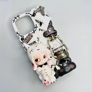 May include: White phone case with a stitched design, featuring a small white cat figurine with a brown hat, a miniature lantern, and a black and white cat head.