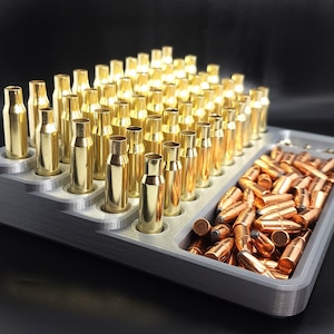 May include: A gray plastic tray with two compartments. The larger compartment holds rows of gold-colored bullet casings. The smaller compartment holds a pile of copper-colored bullets.