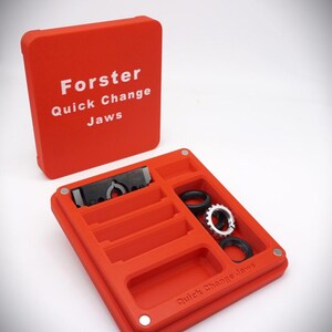 May include: An open, bright red case with the text "Forster Quick Change Jaws" in white. The case contains several compartments holding black and silver metal components. The lid is open, revealing the interior. The text "NOT Included" is above the case.
