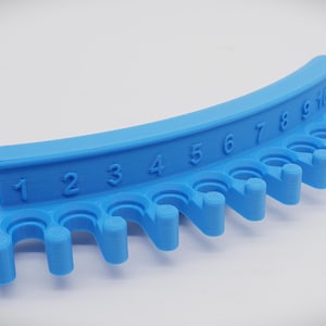 May include: A blue plastic knitting needle holder with 10 numbered pegs. The holder is curved and has a handle on the left side.